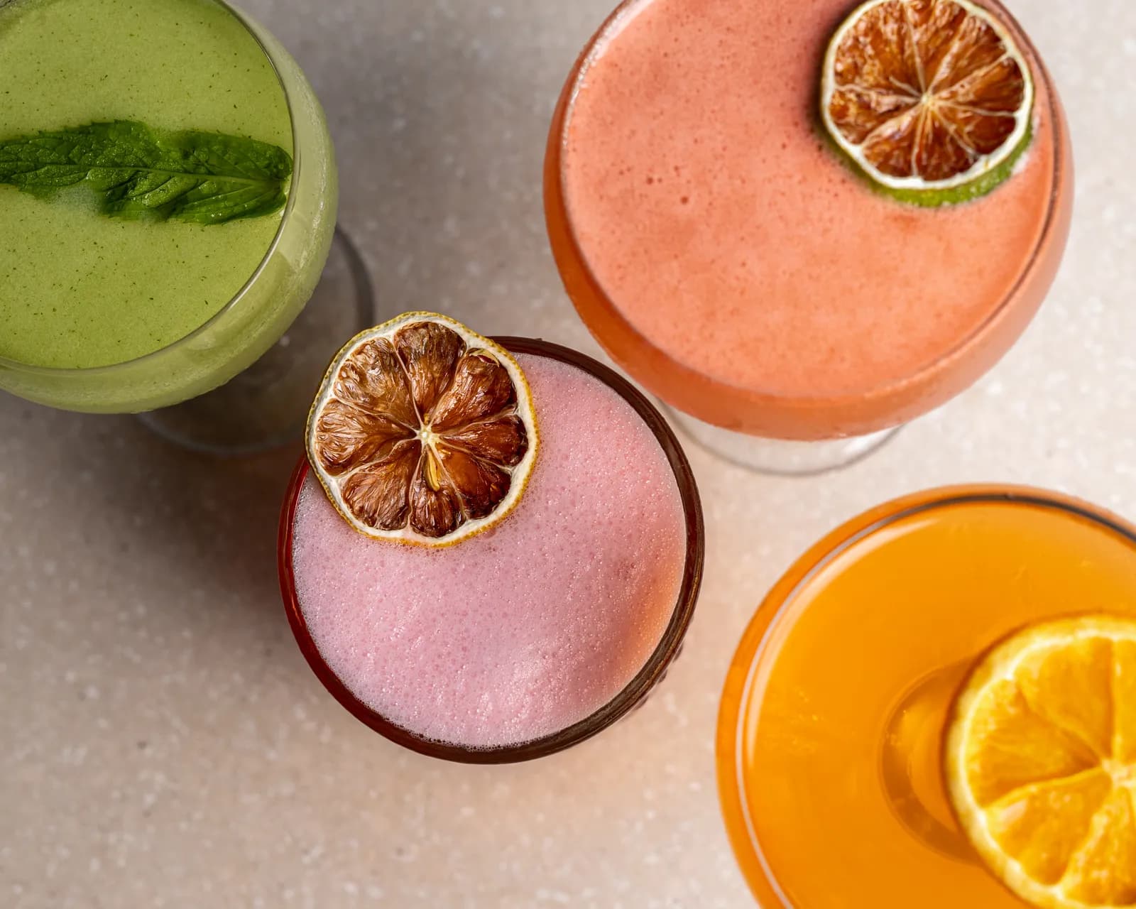 Four cocktails shot from above — green with mint, coral, pale pink and orange, each with a dried citrus wheel